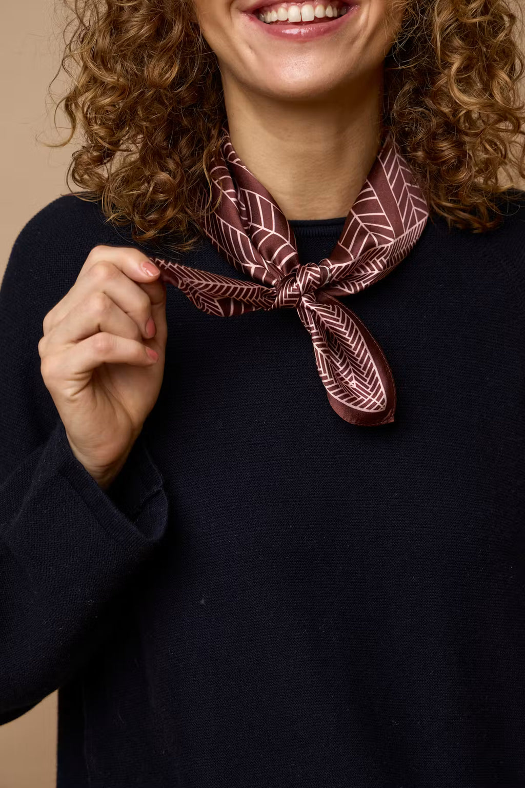 A close-up of a person wearing a dark navy knit sweater and a patterned Rum Raisin  tied around the neck. The scarf features a geometric herringbone design in cream and burgundy.