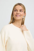 A woman with blonde hair wearing a Whisper White pullover, styled with silver geometric stud earrings and a silver ring, set against a plain light gray background.