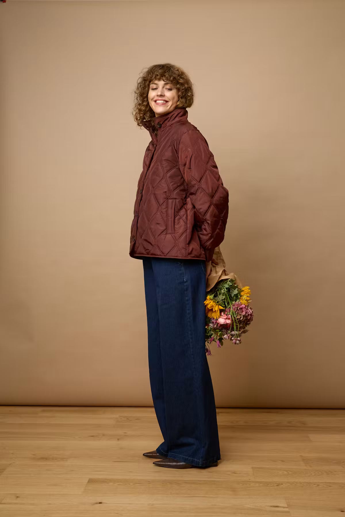 A woman stands in profile wearing a FRAGATE Outerwear in Rum Raisin/Red and wide-leg navy trousers, holding a bouquet of flowers against a plain beige studio backdrop with a light wood floor.