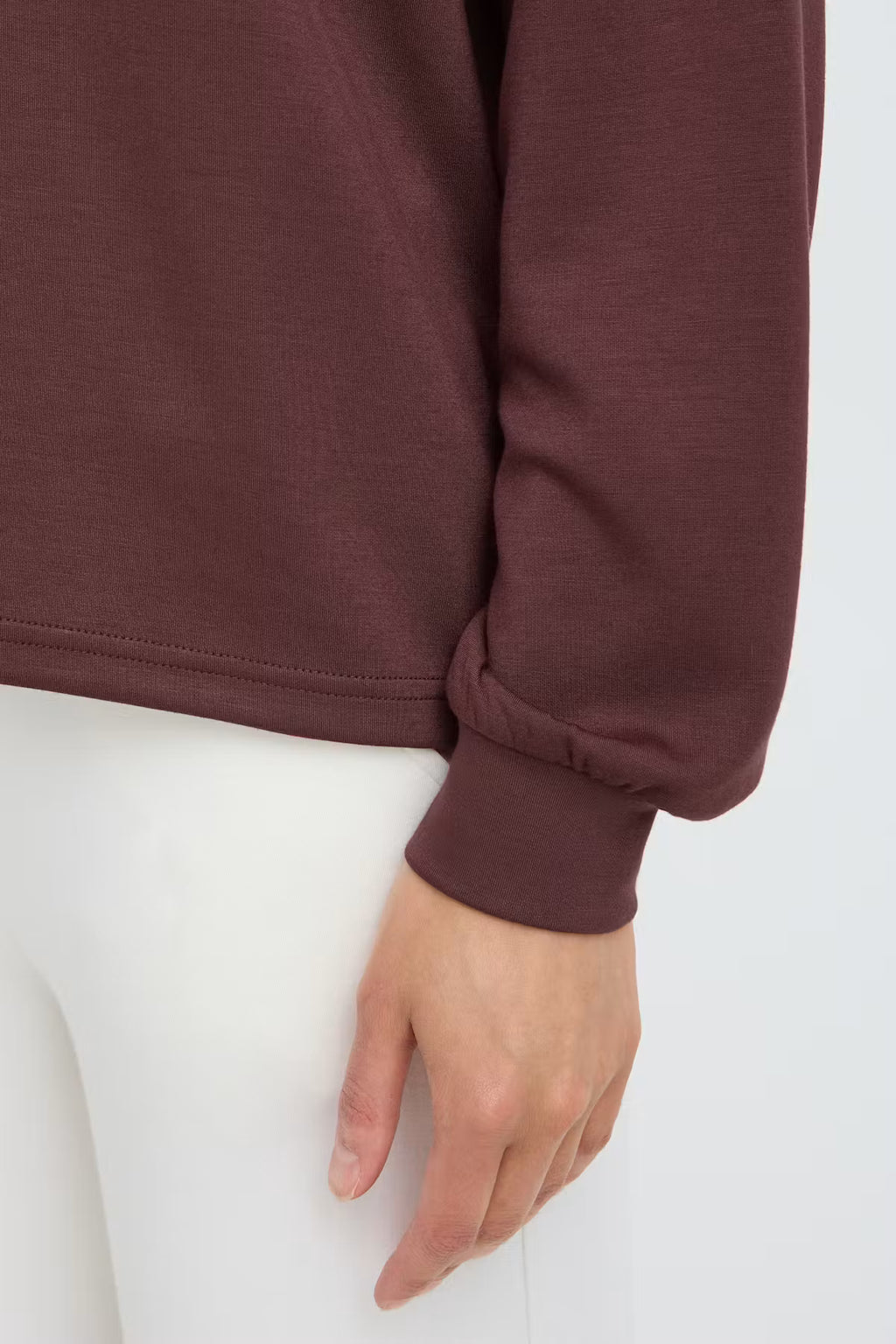 Close-up of a person FRAURORA PU 7 Sweatshirt in Rum Raisin/Red  paired with white pants against a plain light background.
