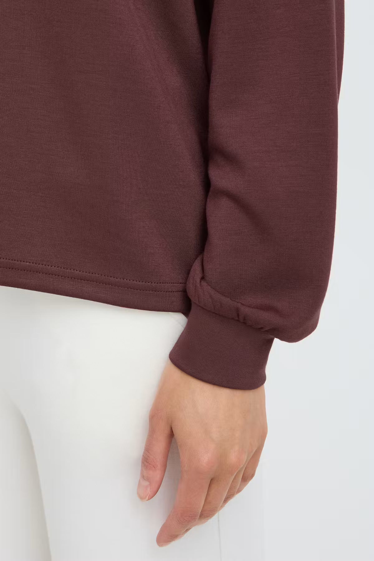 Close-up of a person FRAURORA PU 7 Sweatshirt in Rum Raisin/Red  paired with white pants against a plain light background.