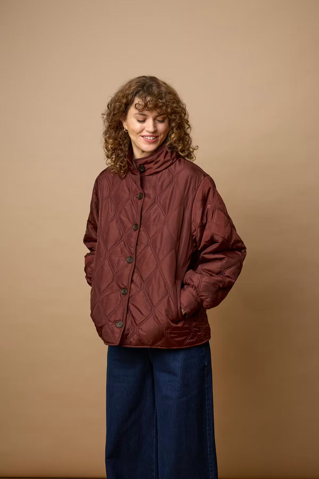 A woman with curly brown hair wearing FRAGATE Outerwear in Rum Raisin/Red, paired with dark blue wide-leg pants, standing against a solid tan studio background.