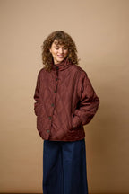 A woman with curly brown hair wearing FRAGATE Outerwear in Rum Raisin/Red, paired with dark blue wide-leg pants, standing against a solid tan studio background.