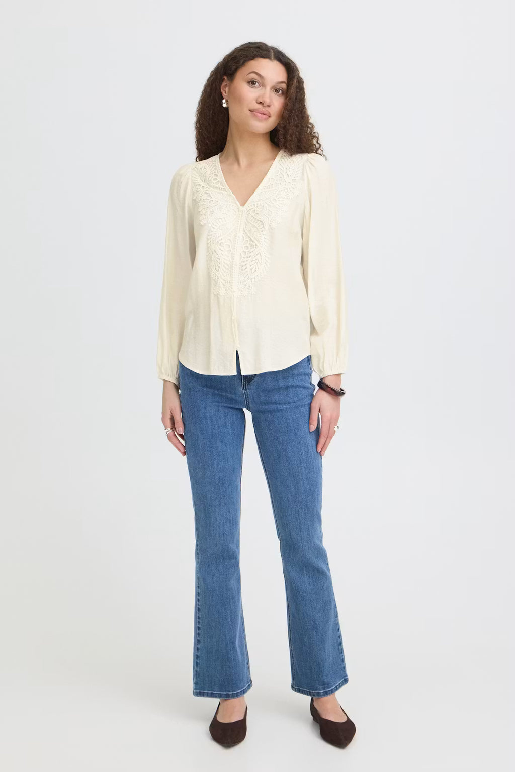A woman modeling a Whisper White long-sleeve blouse with delicate lace embroidery on the front, paired with blue flare jeans and dark brown flats, against a plain light gray studio background.