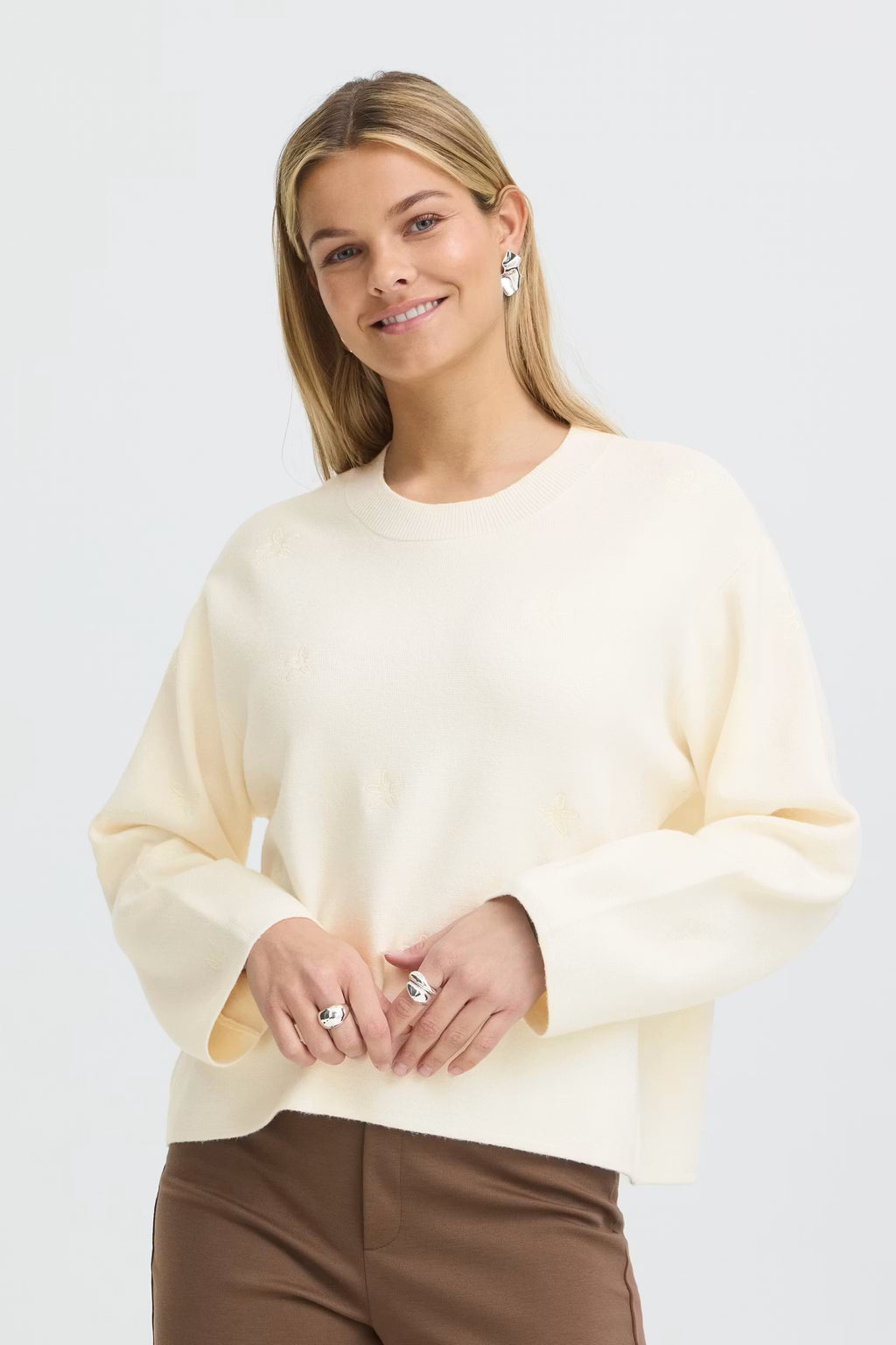 A woman wearing a Whisper White long-sleeve knit sweater and brown trousers, standing against a light gray studio background.