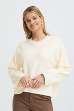 A woman wearing a Whisper White long-sleeve knit sweater and brown trousers, standing against a light gray studio background.