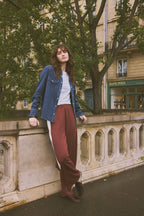 A woman stands outdoors on a stone bridge in Paris, wearing a blue denim shirt over a white top and maroon wide-leg trousers with a white side stripe. Trees and a building are visible in the background.