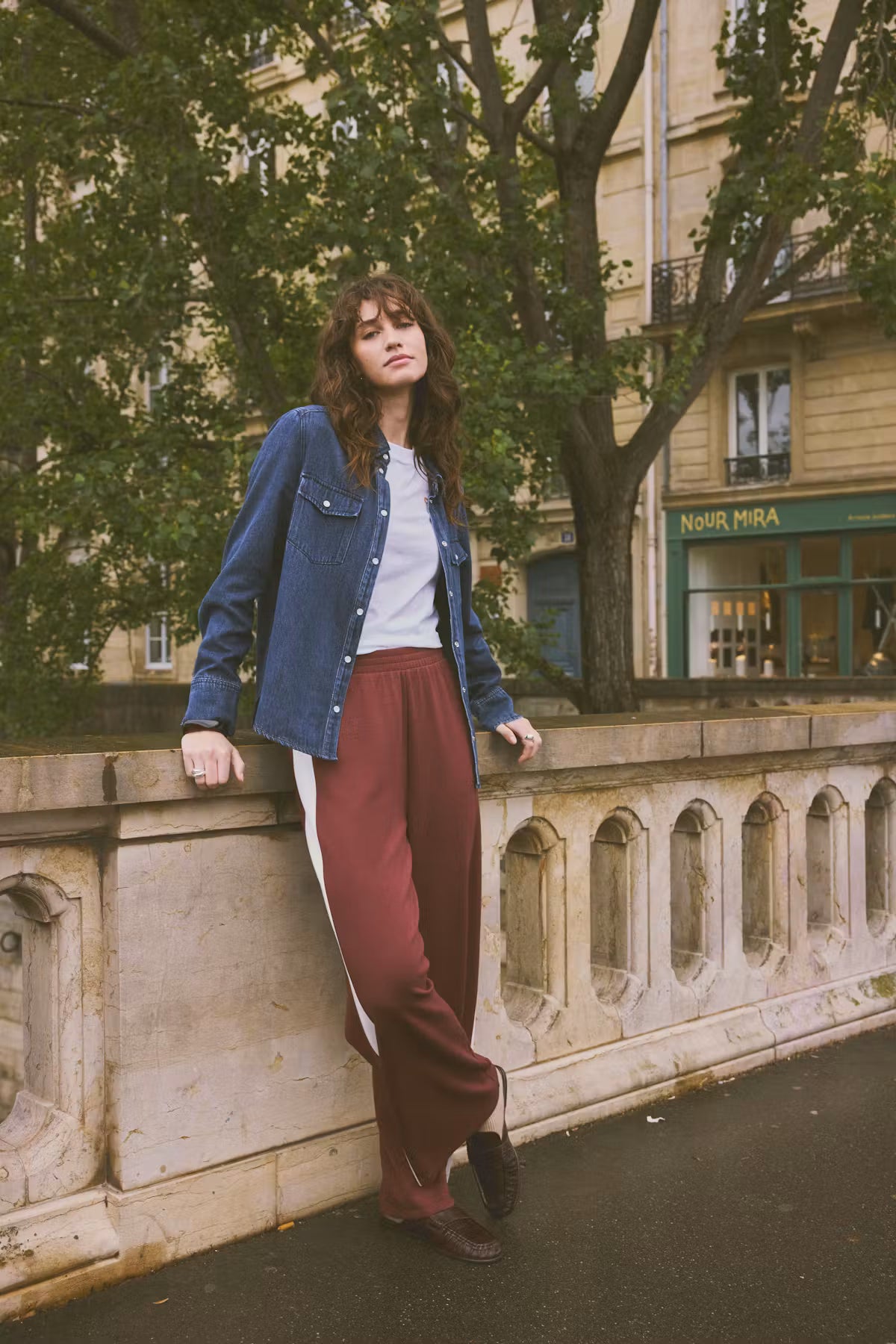 A woman stands outdoors on a stone bridge in Paris, wearing a blue denim shirt over a white top and maroon wide-leg trousers with a white side stripe. Trees and a building are visible in the background.