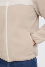 Close-up of a FRFLEA Outerwear in String/Nature with a white contrast panel across the upper chest and white zipper trim, shown on a model with blue jeans against a light gray background.