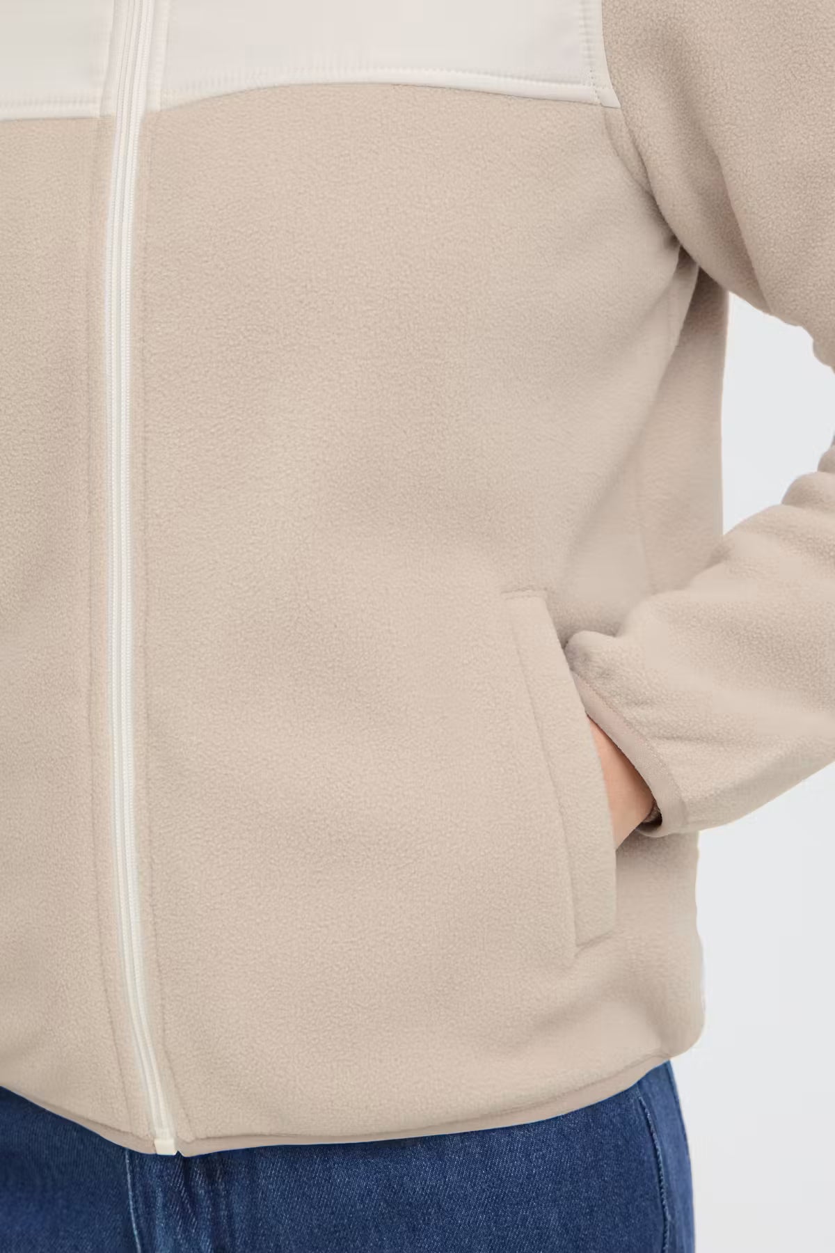 Close-up of a FRFLEA Outerwear in String/Nature with a white contrast panel across the upper chest and white zipper trim, shown on a model with blue jeans against a light gray background.