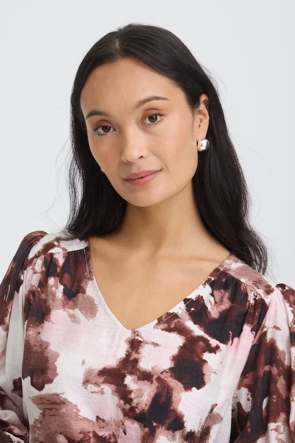 A woman with long dark hair wearing a Watercolour Bloom Rum Raisin / Red V-neck blouse photographed against a light gray studio background.