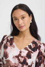 A woman with long dark hair wearing a Watercolour Bloom Rum Raisin / Red V-neck blouse photographed against a light gray studio background.