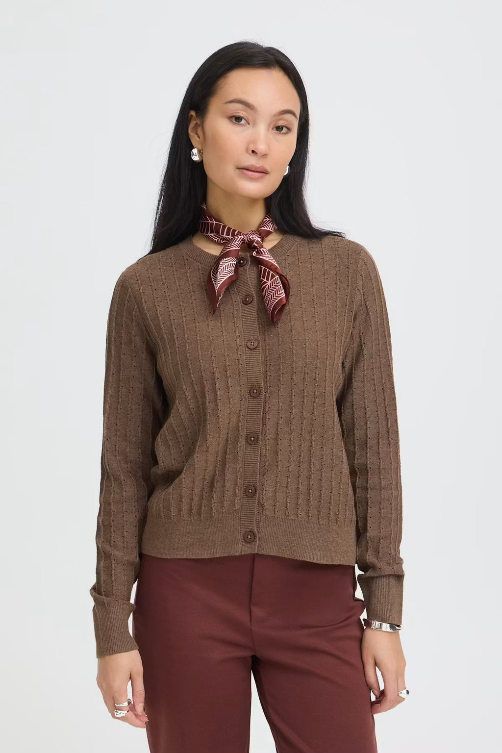 A woman modeling a brown ribbed button-front cardigan sweater with a Rum Raisin neck scarf, paired with burgundy trousers, against a light gray studio background.