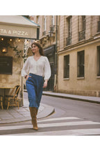 A woman walking across a city street crosswalk wearing a Whisper White long-sleeve blouse with a V-neck, blue high-waisted wide-leg trousers, and brown knee-high boots. The background shows a European-style street with beige buildings and a pizza shop.