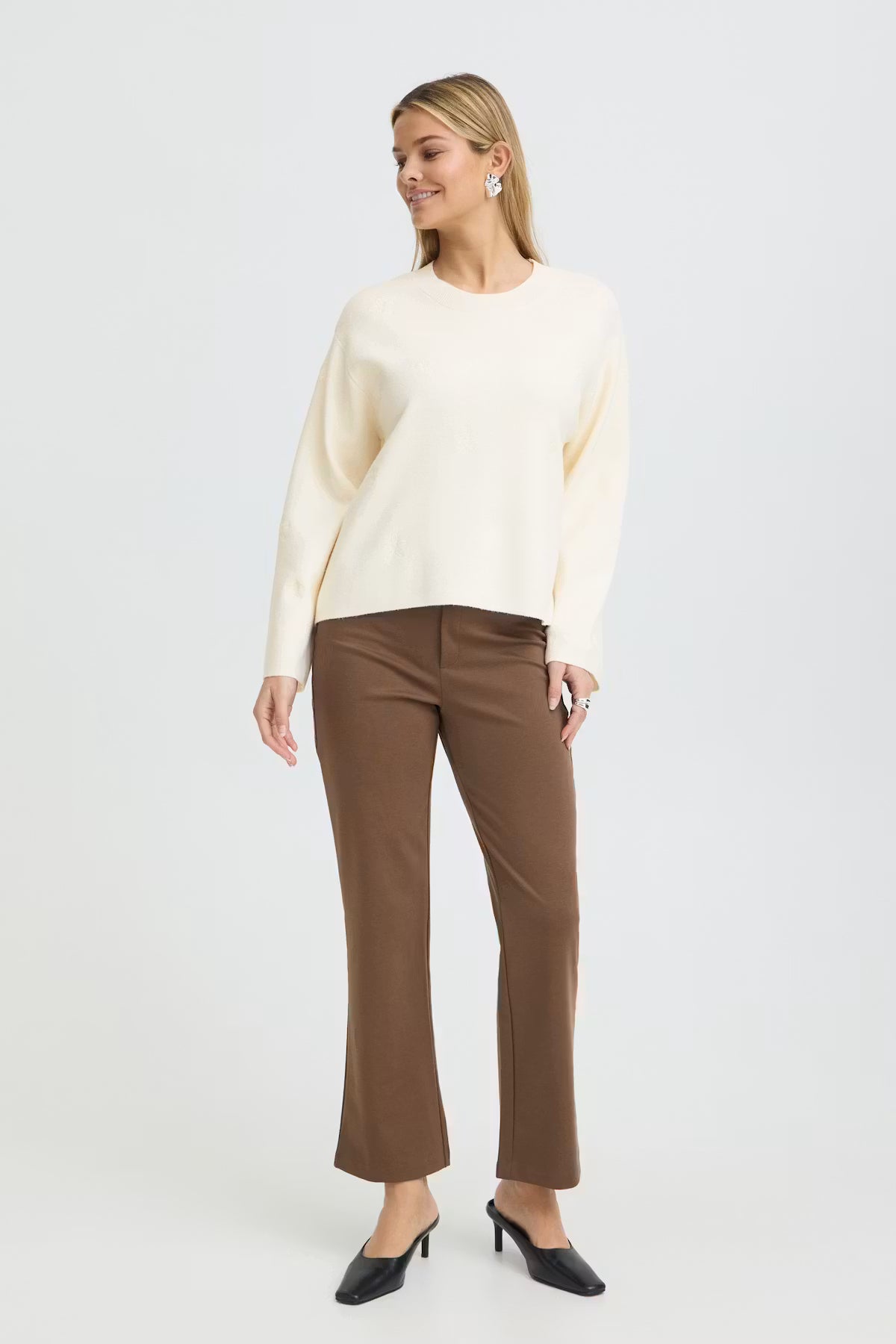 A woman modeling a Whisper White pullover and brown cropped trousers with black pointed-toe heels, set against a plain light gray studio background.