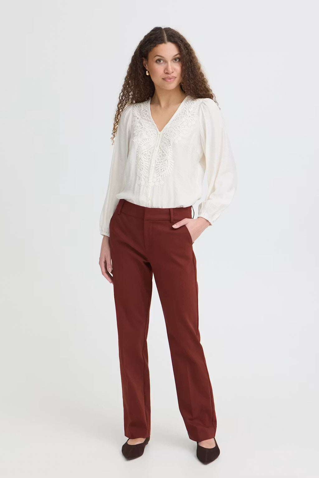 A woman modeling a white long-sleeve blouse with lace detailing on the chest and dark rust-brown straight-leg 
Rum Raisin / Red trousers, standing against a plain light gray studio background.