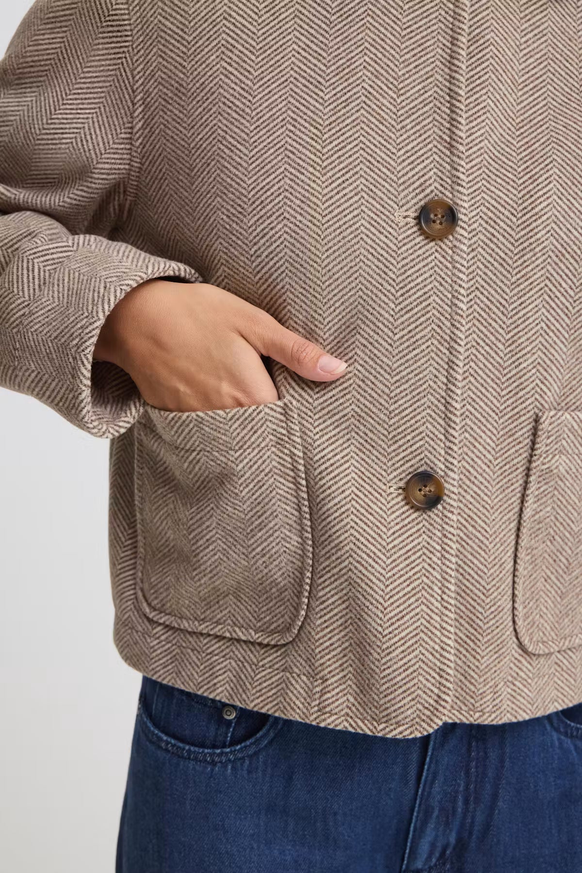 Close-up of a person wearing a FREVILLA Outerwear in Rain-Drum  Mix/Brown with dark buttons and front patch pockets, paired with blue jeans against a light gray background.