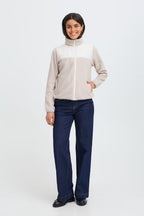 A woman modeling a FRFLEA Outerwear in String/Nature with a high mock neck, featuring beige and white panels, paired with dark blue jeans and black shoes, against a plain white studio background.