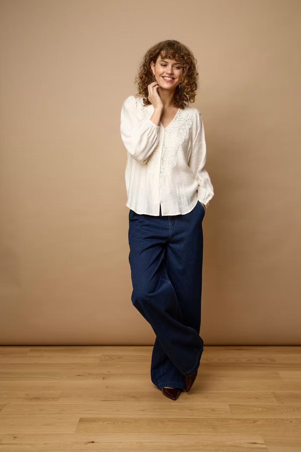 A woman with curly brown hair stands in a studio wearing a Whisper White long-sleeve blouse with lace detailing and dark navy wide-leg trousers, posed against a solid beige backdrop and light wood floor.