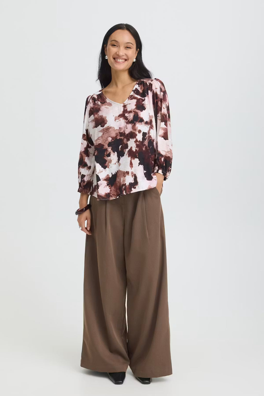 A woman modeling a Watercolour Bloom Rum Raisin / Red long-sleeve blouse paired with wide-leg brown trousers, against a light gray studio background.