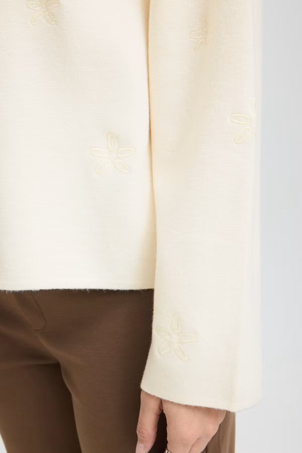 Close-up of a Whisper White pullover with subtle tonal floral embroidery on the sleeve, worn with brown trousers.