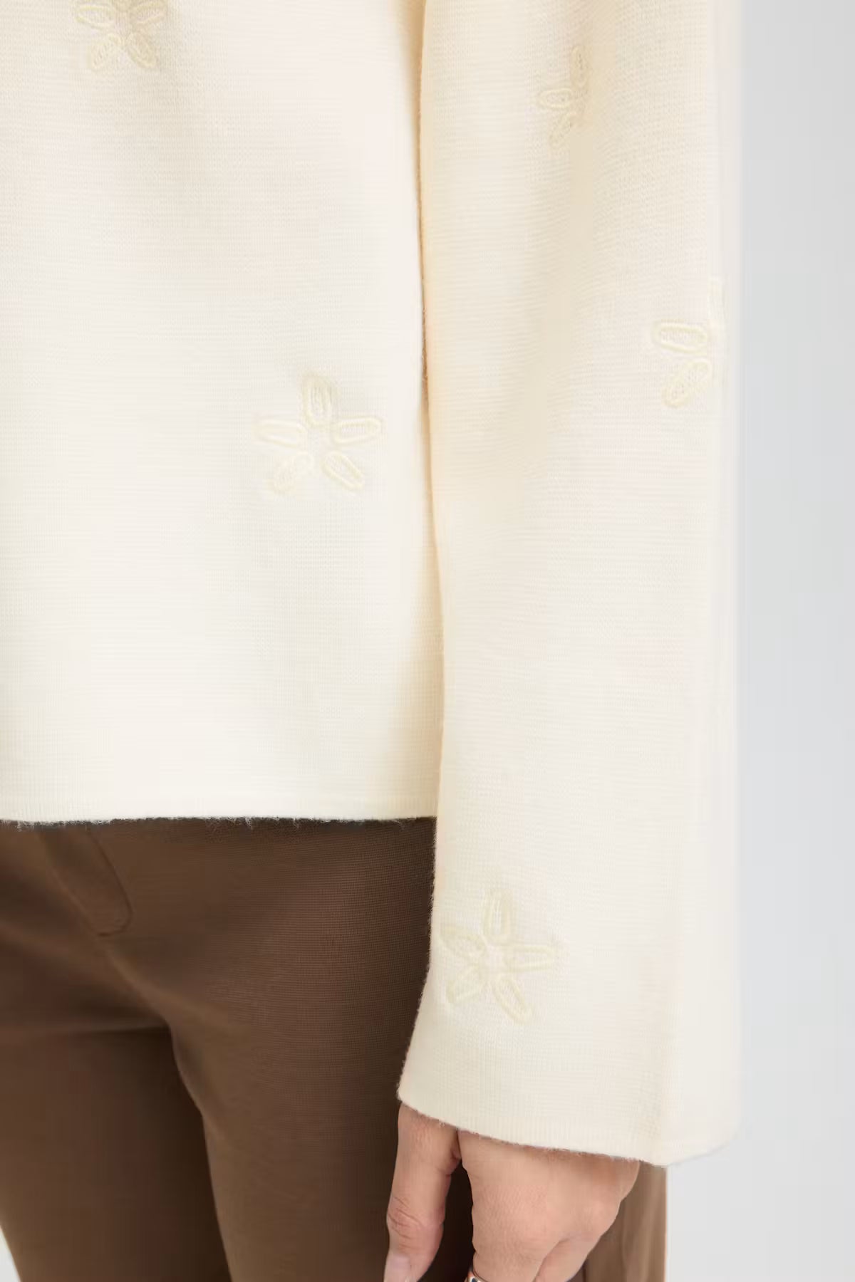 Close-up of a Whisper White pullover with subtle tonal floral embroidery on the sleeve, worn with brown trousers.