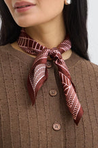 A close-up of a woman wearing a brown ribbed knit cardigan sweater and a patterned Rum Raisin  scarf tied around her collar. The scarf features a geometric chevron design in dark red and white.