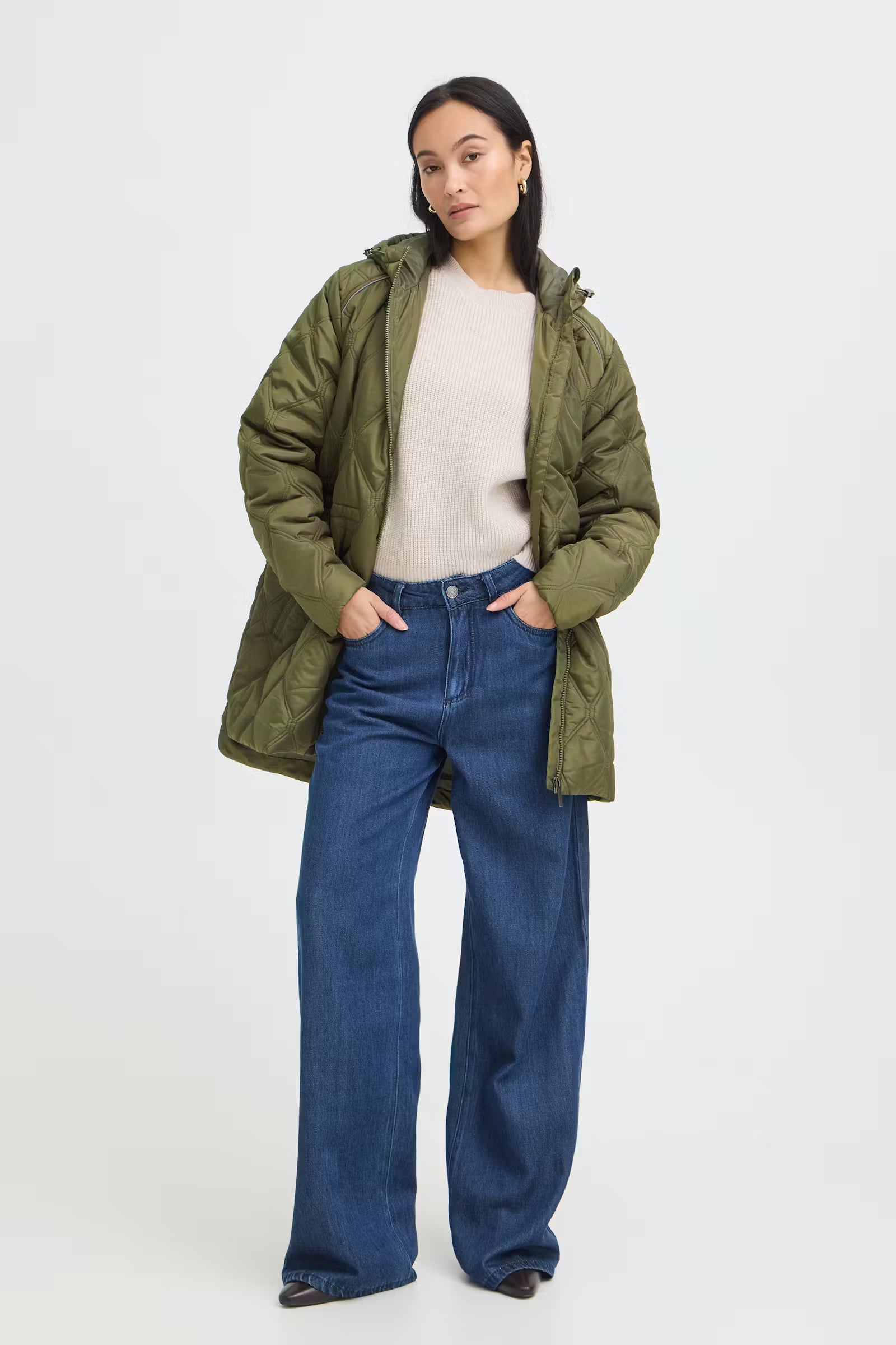 A woman stands against a light gray studio background wearing aFRAGATE Outerwear in Grape Leaf/Green puffer jacket over a cream ribbed top and blue wide-leg jeans.