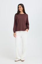 A woman modeling FRAURORA PU 7 Sweatshirt in Rum Raisin/Red , paired with white wide-leg pants and white shoes, against a plain light gray studio background.
