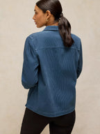 A woman shown from the back wearing a blue corduroy button-up shirt and black pants, standing against a textured beige wall.