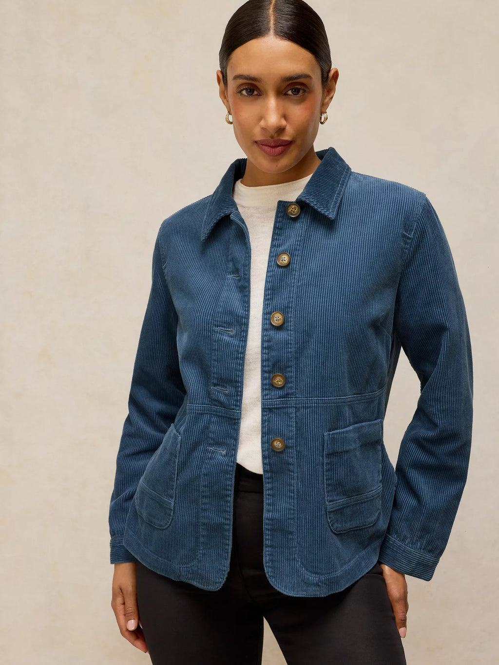 A woman wearing a blue corduroy jacket with brown buttons over a white top and black pants, standing against a textured beige background.