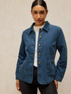 A woman wearing a blue corduroy jacket with brown buttons over a white top and black pants, standing against a textured beige background.