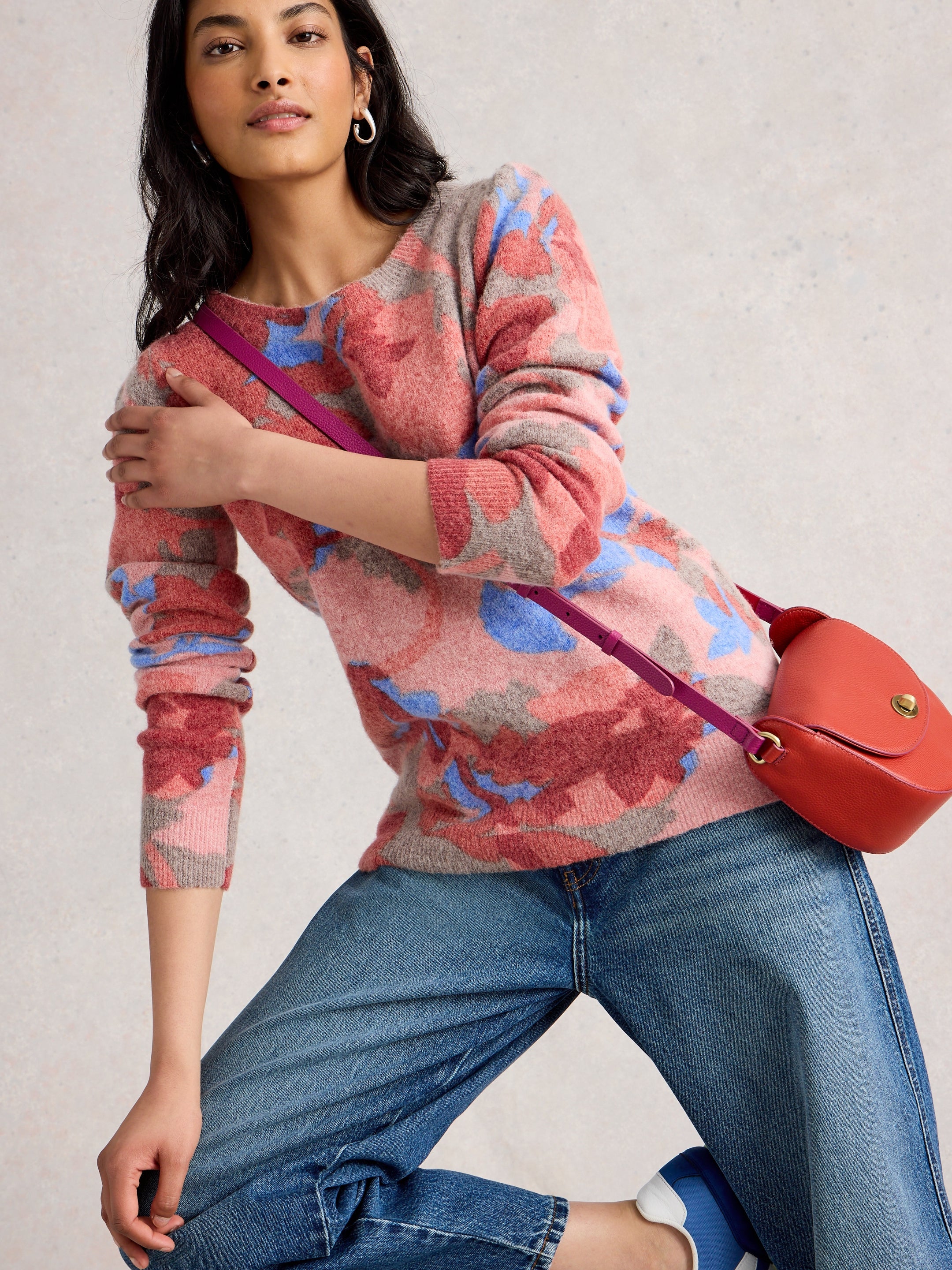 Woman wearing a colorful sweater and jeans, holding a red bag against a neutral background