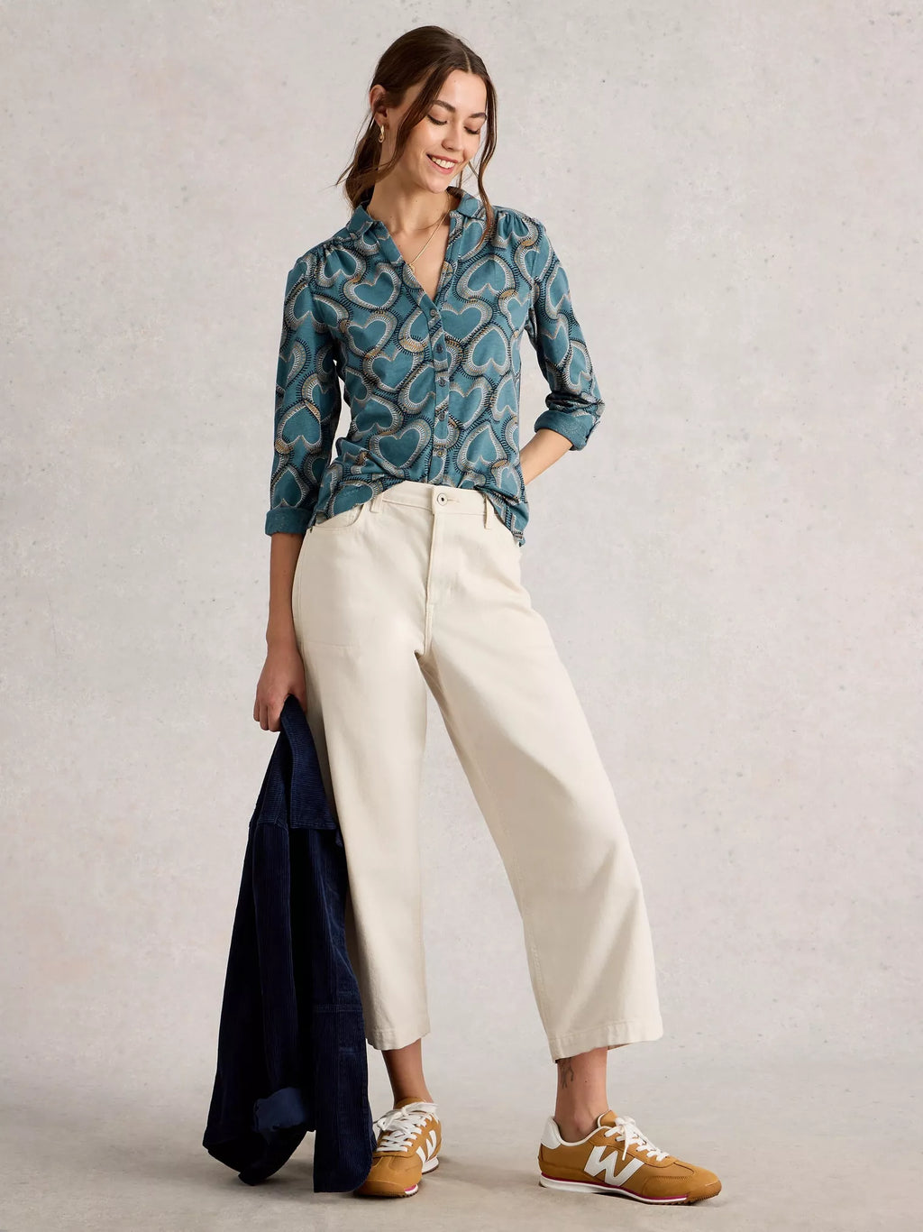 Woman wearing a blue patterned shirt and white pants, holding a dark jacket against a light background