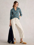 Woman wearing a blue patterned shirt and white pants, holding a dark jacket against a light background