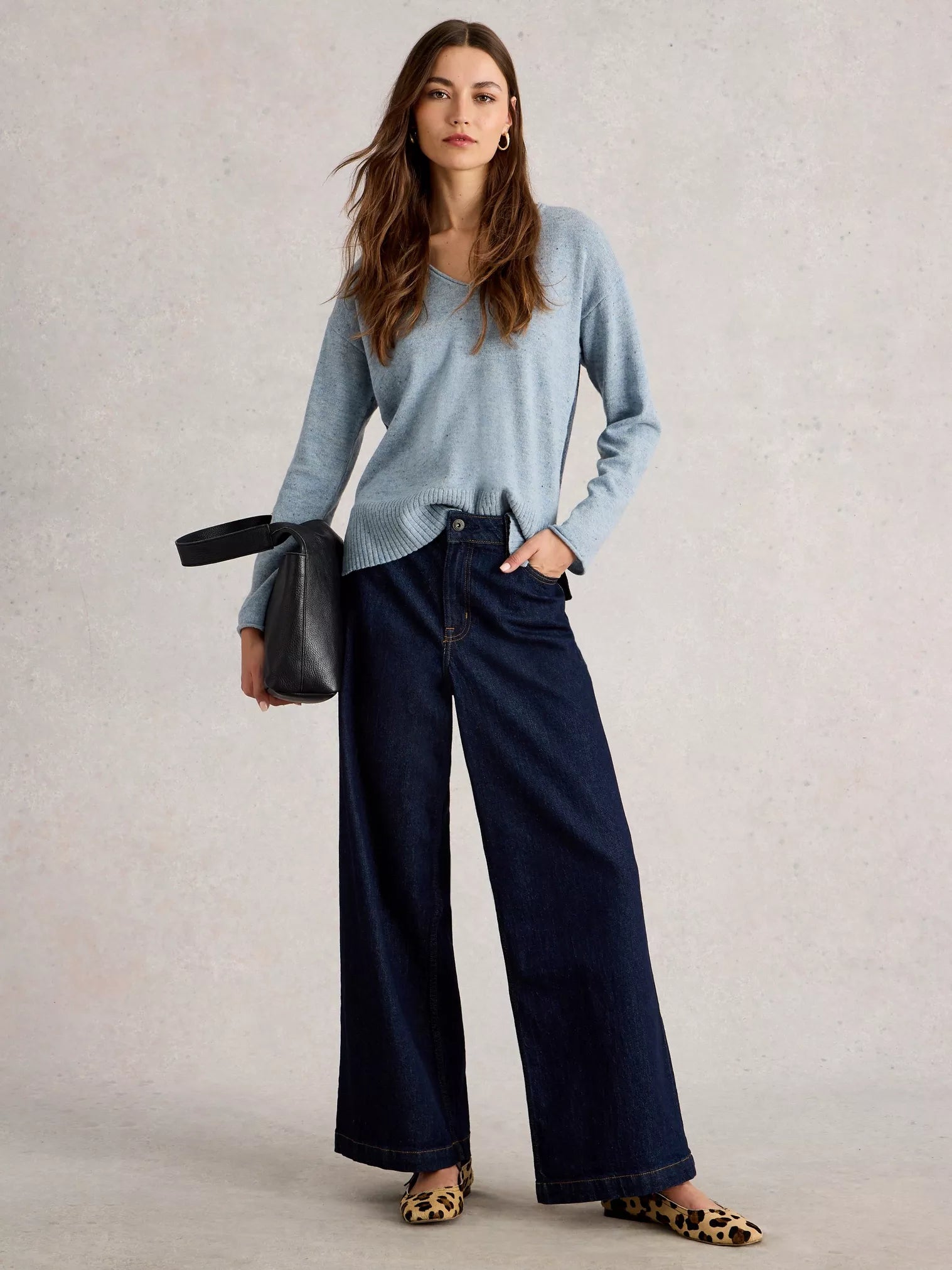 Woman wearing a light blue sweater and dark blue jeans, holding a black handbag on a plain background