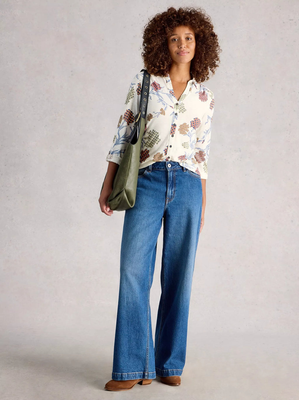 Woman wearing a floral blouse and blue jeans, holding a green bag against a plain background