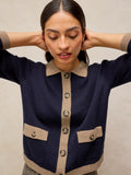 A woman wearing a navy blue knit cardigan with beige trim, a beige collar, and beige button placket, posing with her hands behind her head against a neutral beige background.