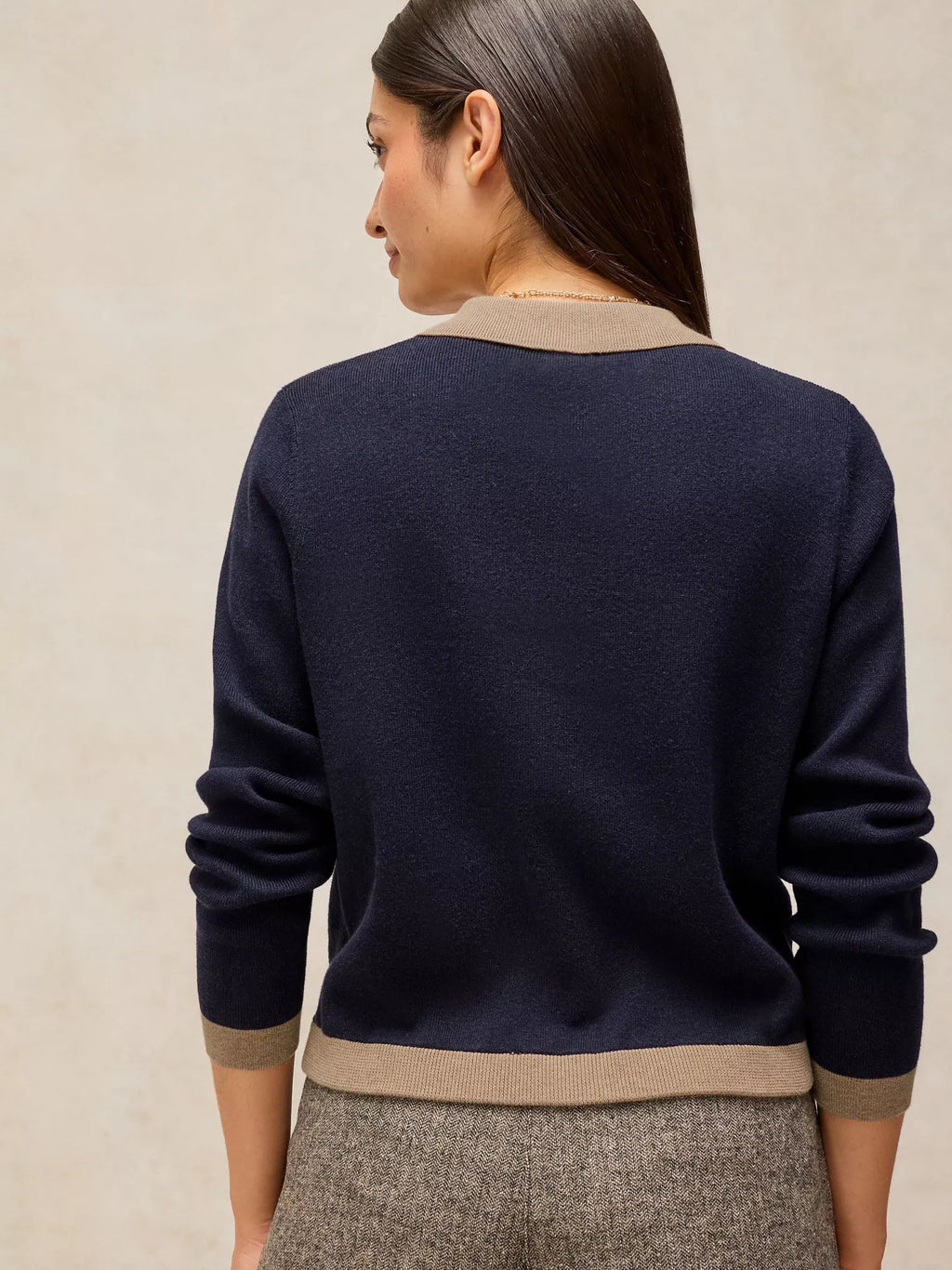 A woman shown from the back wearing a navy blue knit sweater with beige ribbed trim at the collar, cuffs, and hem, paired with gray textured trousers against a neutral beige background.