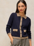 A woman wearing a navy blue knit cardigan with beige trim, featuring a pointed collar, button-front closure, and flap pockets, paired with gray textured trousers against a neutral beige background.