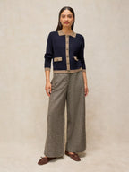 A woman stands against a warm beige studio backdrop wearing a navy cardigan with beige trim and buttons, paired with wide-leg gray trousers and brown moccasin-style shoes.