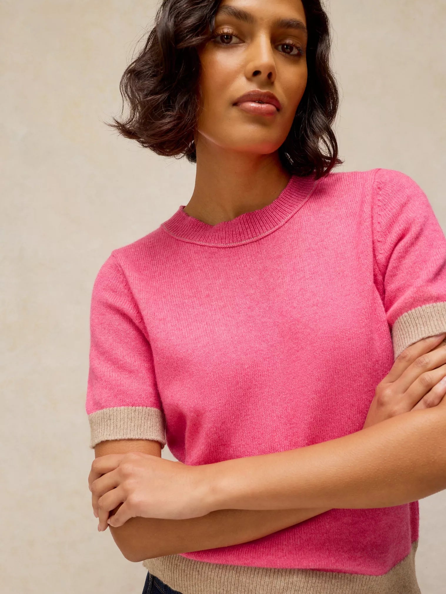 A woman wearing a bright pink short-sleeve knit sweater with beige ribbed trim at the sleeve cuffs and hem, posed against a neutral beige background.