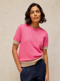 A woman wearing a bright pink short-sleeve knit sweater with beige ribbed trim at the sleeve cuffs and hem, paired with dark blue jeans, against a neutral textured beige background.