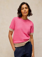 A woman wearing a bright pink short-sleeve knit sweater with beige ribbed trim at the sleeve cuffs and hem, paired with dark blue jeans, against a neutral textured beige background.