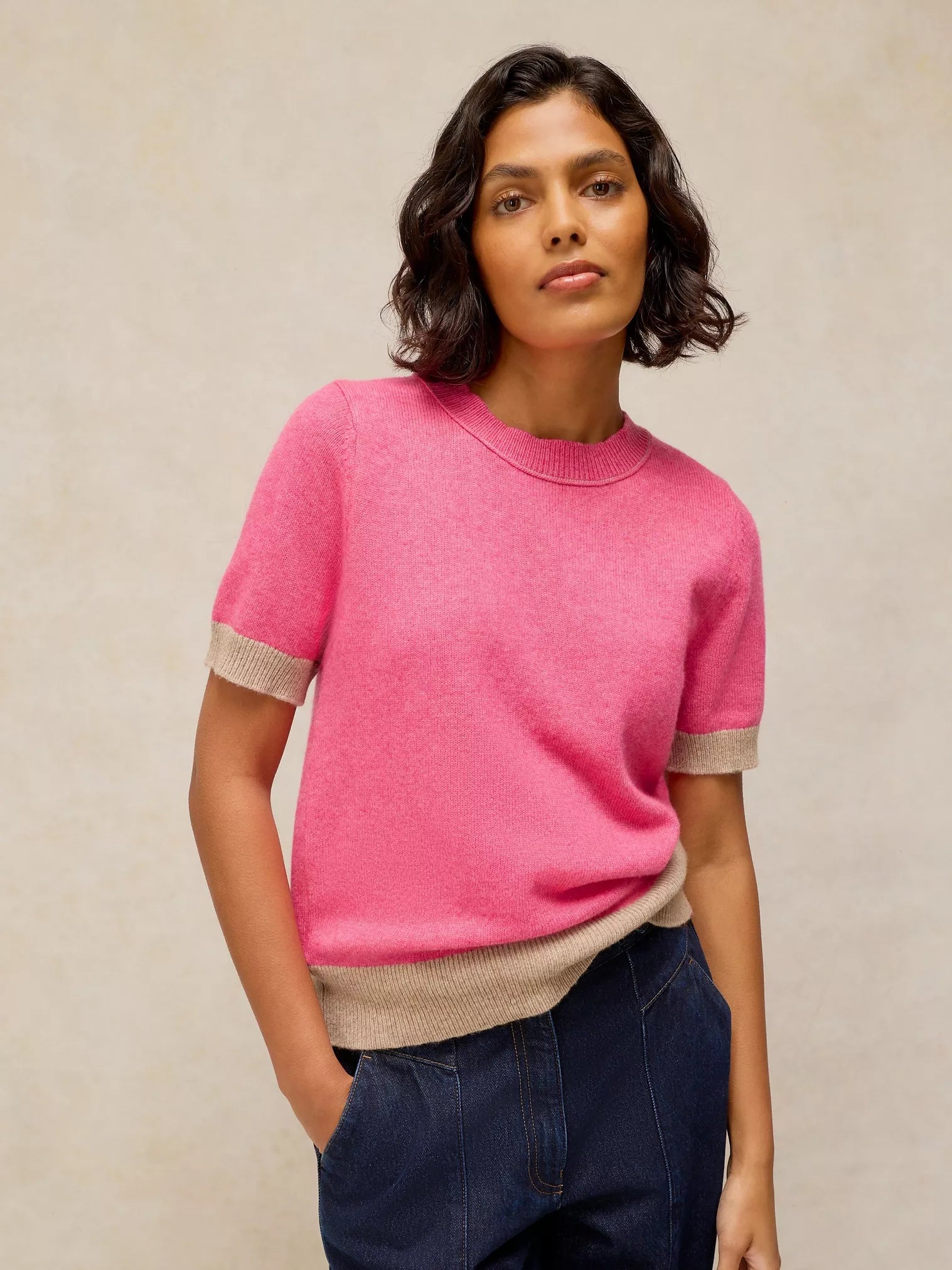 A woman wearing a bright pink short-sleeve knit sweater with beige ribbed trim at the sleeve cuffs and hem, paired with dark blue jeans, against a neutral textured beige background.