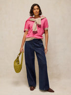 A model stands against a textured beige studio backdrop wearing a bright pink knit top with a beige and burgundy scarf, dark wide-leg jeans, and brown shoes, while holding a yellow-green slouchy shoulder bag.