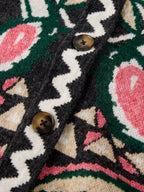 Close-up of a knit cardigan featuring a bold geometric and floral pattern in black, green, cream, and pink, with two dark brown buttons on the front.