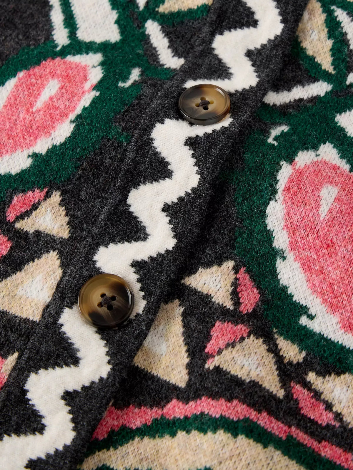 Close-up of a knit cardigan featuring a bold geometric and floral pattern in black, green, cream, and pink, with two dark brown buttons on the front.