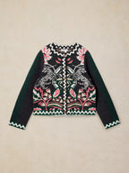 A cropped knit cardigan sweater featuring a black base with a colorful embroidered pattern of birds, leaves, and geometric motifs, with solid dark green sleeves and zigzag trim, laid flat on a beige background.