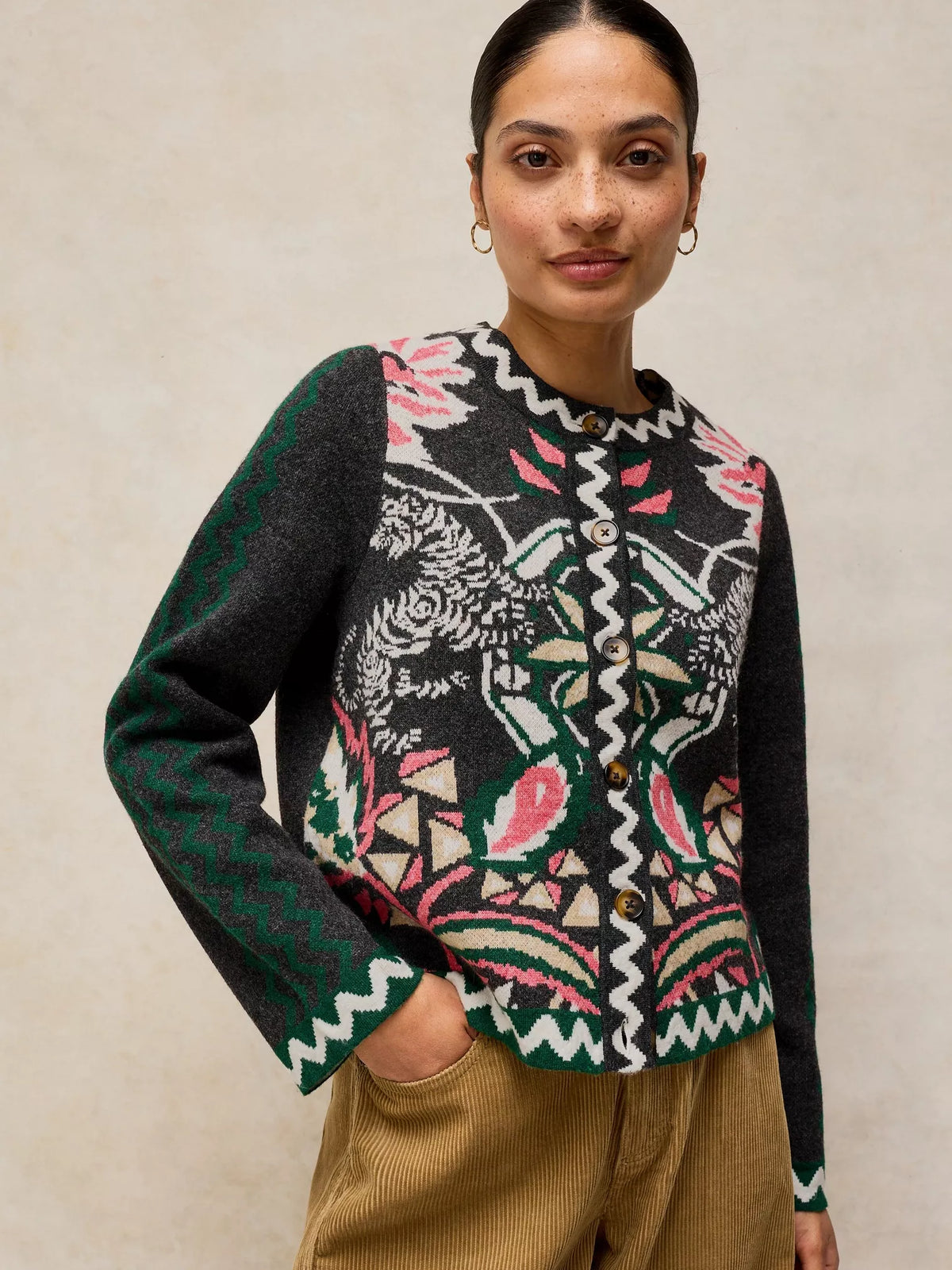 A woman wearing a cropped button-front cardigan with a black base and a colorful floral and geometric pattern in pink, green, white, and beige, paired with tan corduroy trousers against a neutral beige background.